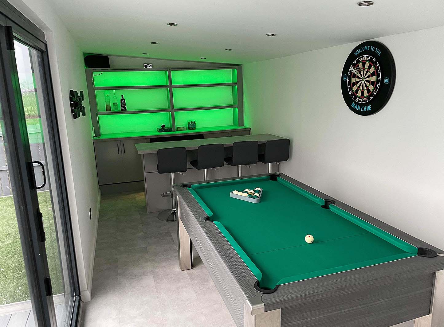 games room in a green theme with a pool table and darts board and bar with dark grey stools