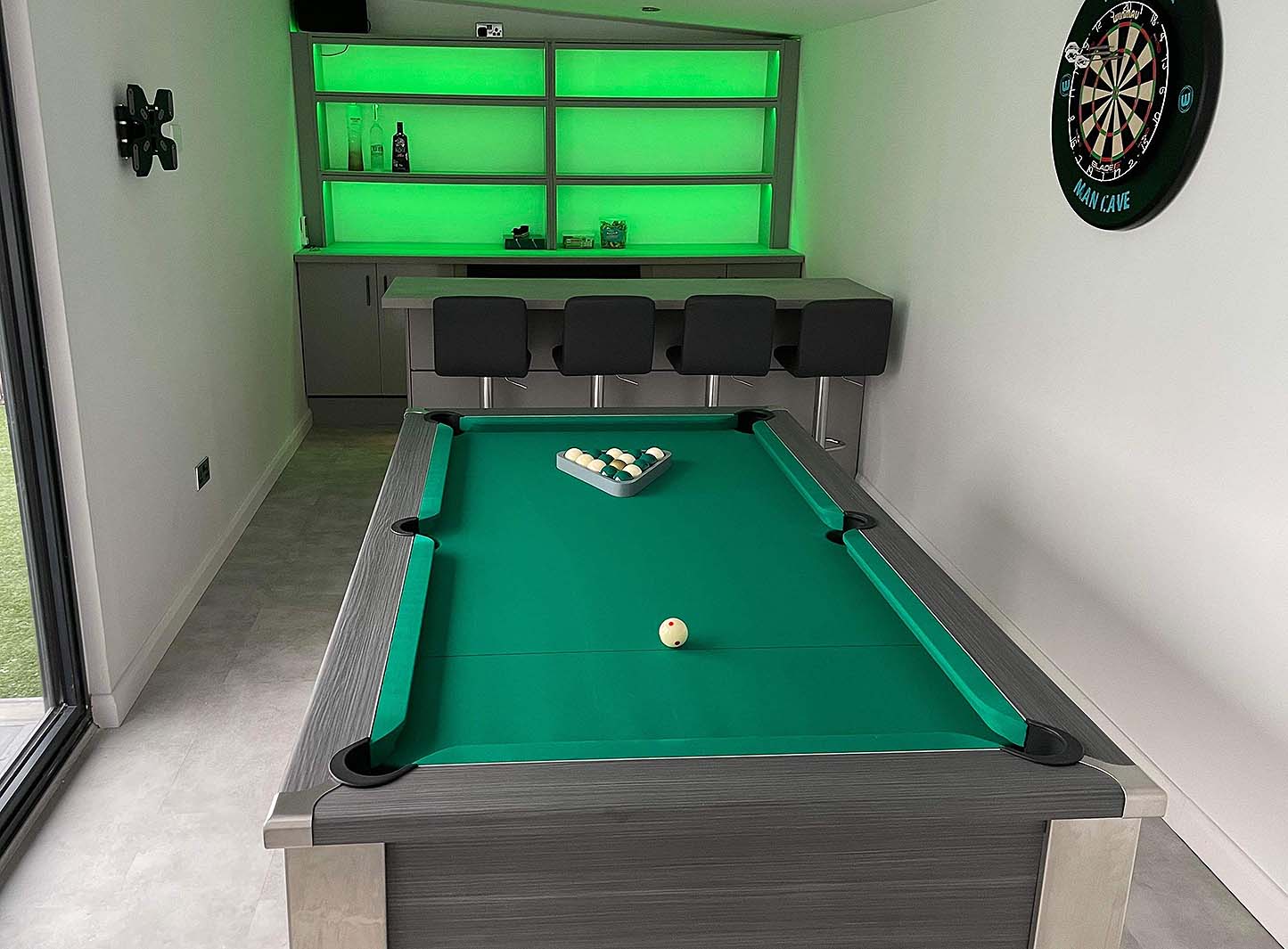 games room in a green theme with a pool table and darts board and bar with dark grey stools